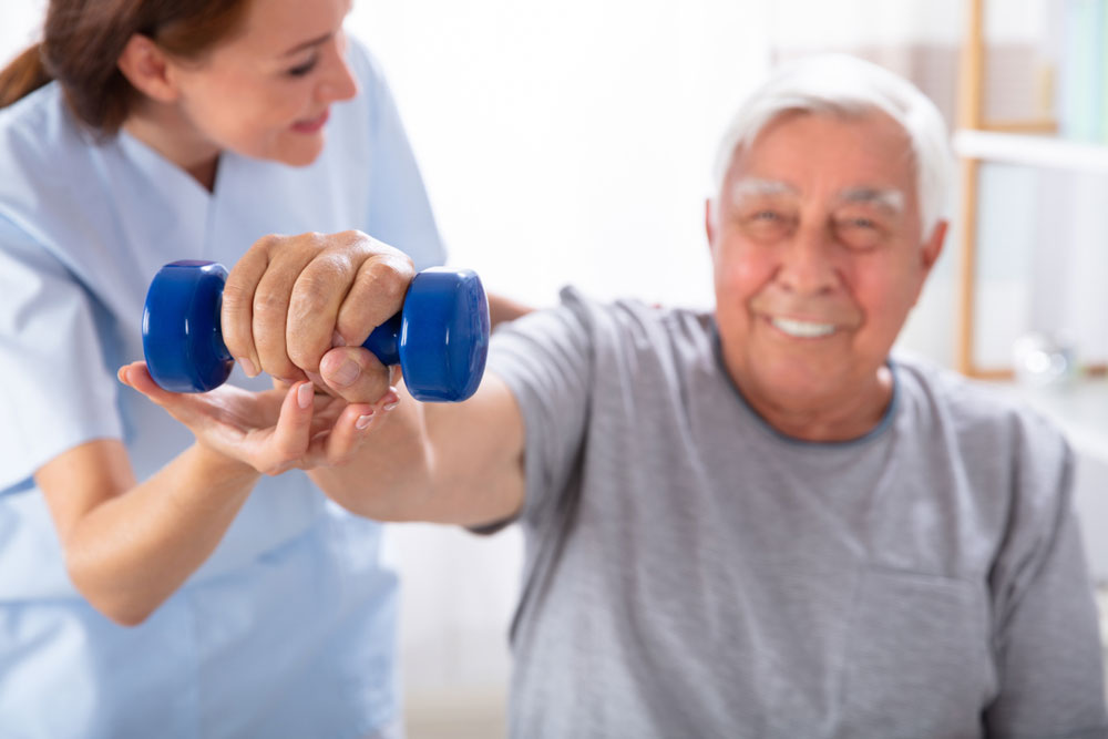 Young Female Nurse Helping Happy Senior Man With Dumbbell Exercise